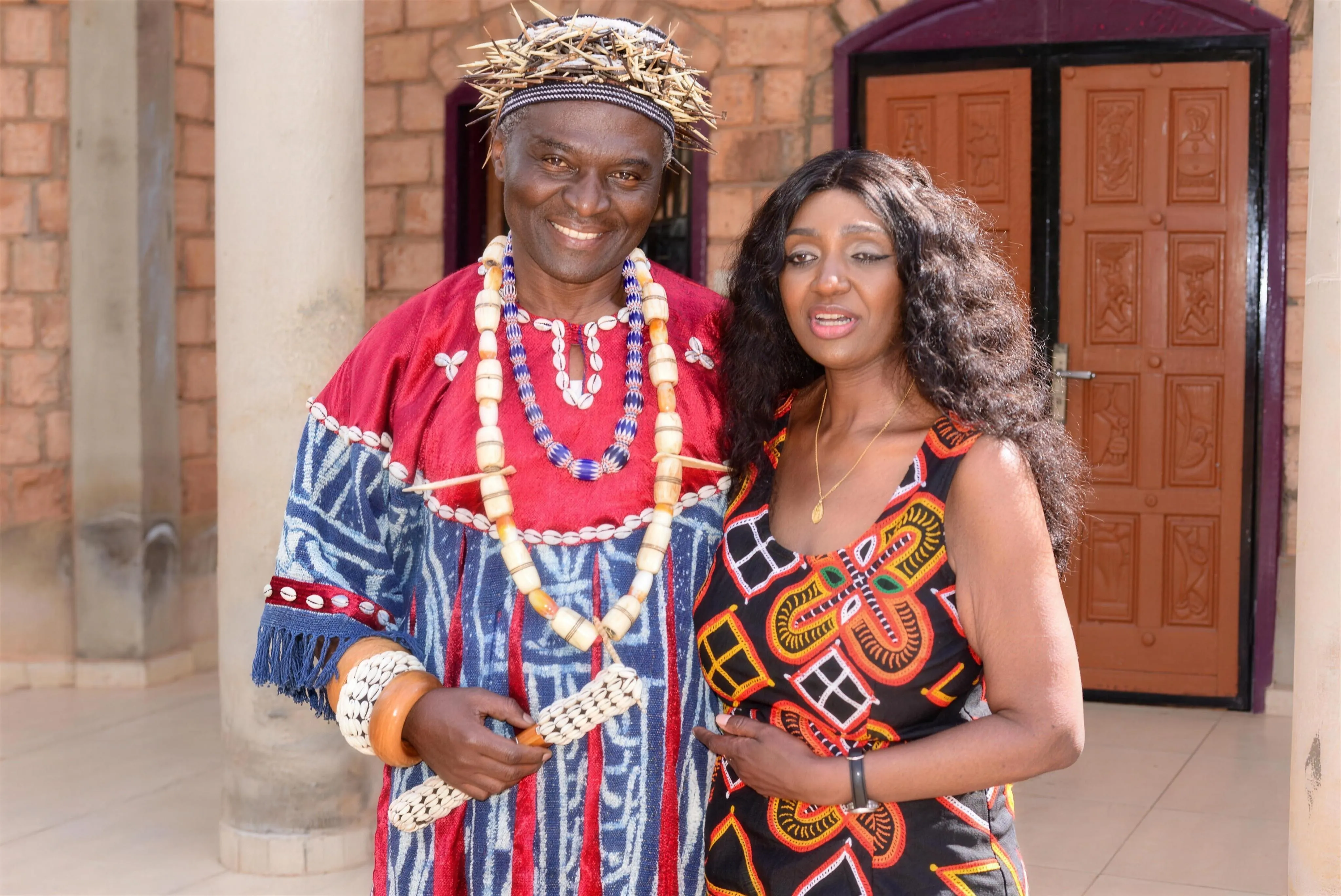Angélina Tezanou aux côtés d'un chef bamiléké en tenue traditionnelle (cauris, coiffe) devant une chefferie, au Cameroun.