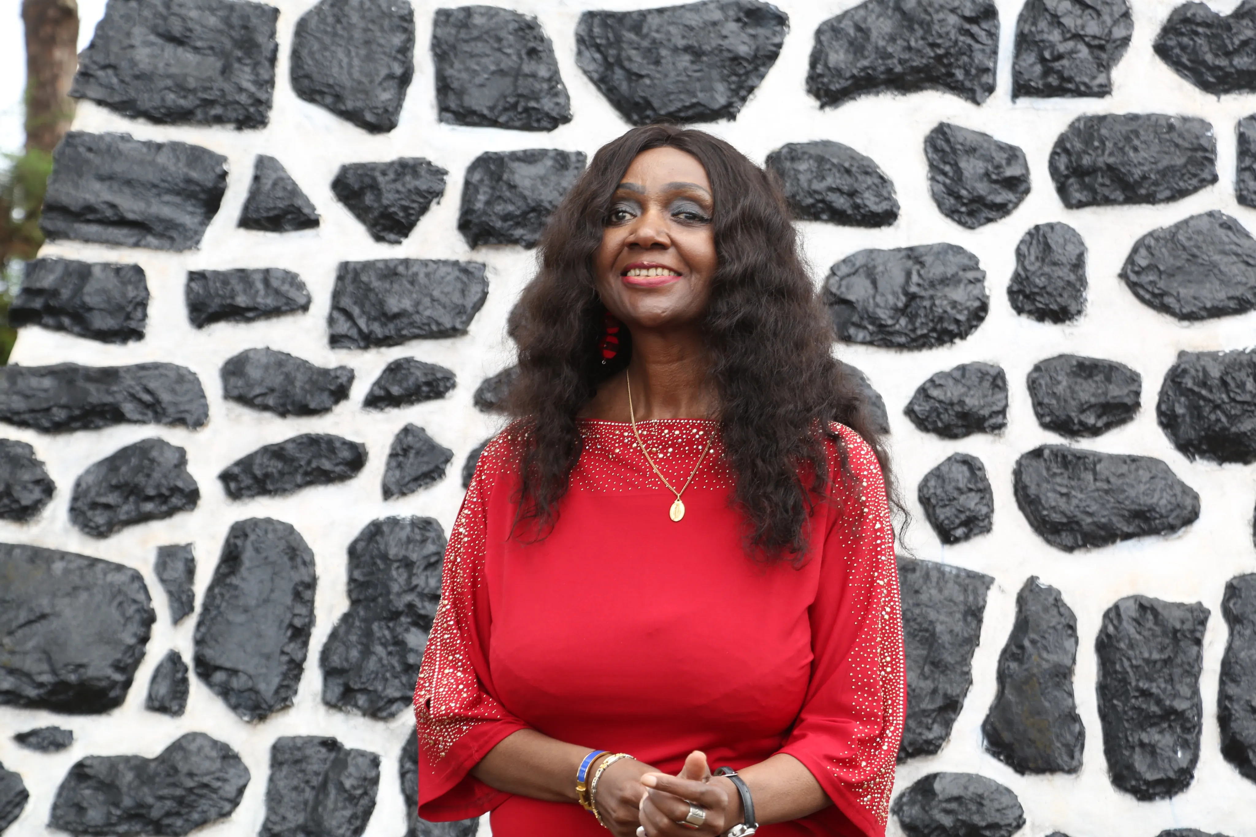 Angélina Tezanou en robe rouge à sequins, mains jointes, devant un mur de pierres de basalte noir et blanc.